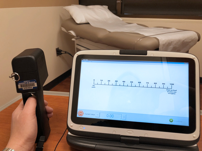 A person’s thumb is wedged in a hole in a hand-held device with a wire coming off it. Next to it is a screen showing a pain scale from 0 to 100.