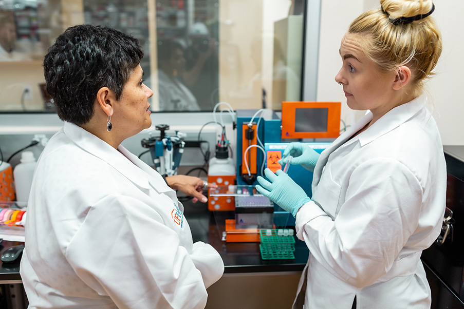 Two women in white lab coats stand in front of a machine that is sitting on a lab bench. The older woman, on the left, is talking to the younger woman, who is wearing blue gloves and holding a small test tube.