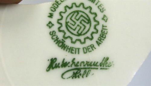 A photograph shows a fragment of a porcelain plate that was excavated at the former Tempelhof airfield in Berlin, site of the old airport and forced labor camps during the Nazi era. The phrase “Schönheit der Arbeit,” inscribed on the plate, means “beauty of work,” and is the name of a Nazi propaganda organization for workplace efficiency. A swastika design is also on the plate.