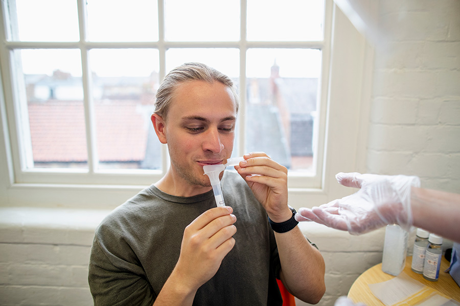 Photograph of a man sitting in a room with white brick walls and in front of a window, spitting into a plastic tube. On the right, an outstretched hand wearing a plastic glove is waiting to be handed the tube with saliva.