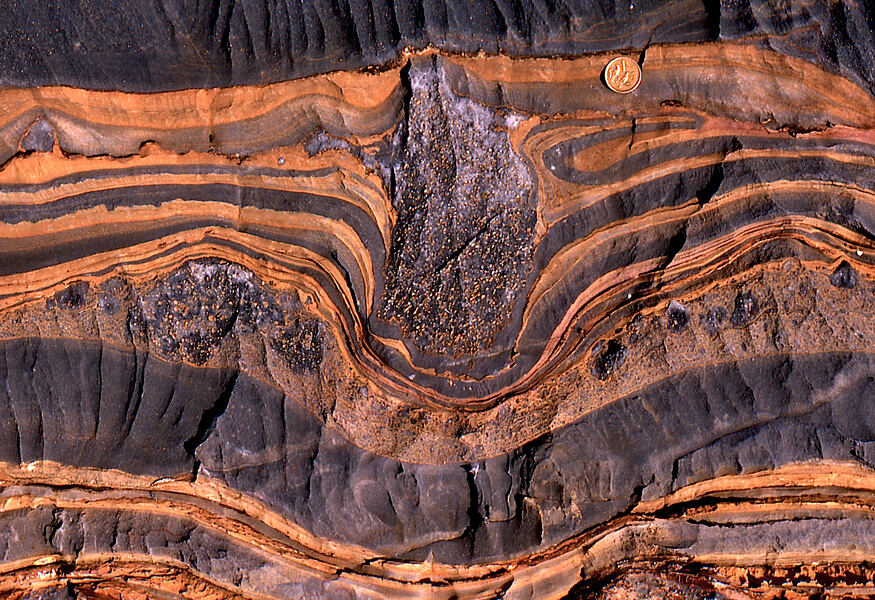 Photograph of a glacial dropstone from Namibia, in rocks that date to the second Snowball Earth. The stone was likely carried and dropped by a floating ice shelf, and when it plunked into seafloor sediment below, that sediment folded around it. (Penny shown for scale.)