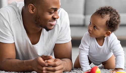 Photo shows a young man smiling at a crawling baby in a living room.