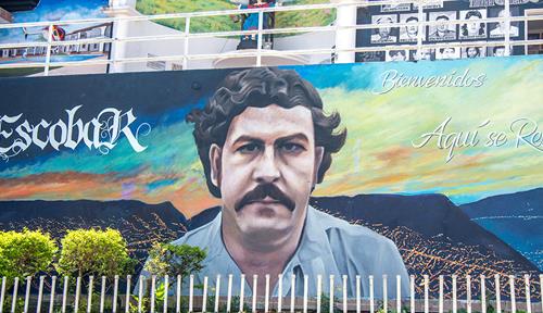 Photo shows a mural of a man with black hair and mustache in front of a landscape with city lights, mountains and the sunset. The words “Barrio Pablo Escobar” are on the left. On the right it says: “Bienvenidos” (Welcome) along with the slogan “Aquí se respira paz” (Here we breathe peace).