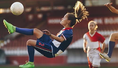 A teenage girl jumps into the air to kick a soccer ball while other players look on.