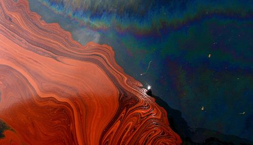 Photograph of an aerial view of the Deepwater Horizon oil spill. The swirls of surface oil are colored red and orange; other parts of the water are blue layered with oily iridescence.