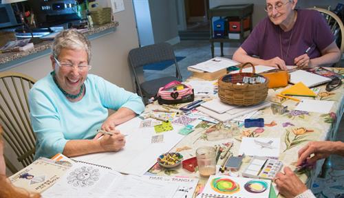 Photo shows a 2016 image of a woman who lives with chronic pain laughing with friends at her dining table while creating Zen tangle art.