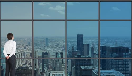 Photo of a man looking out on a city through a large set of windows tinted from dark to clear