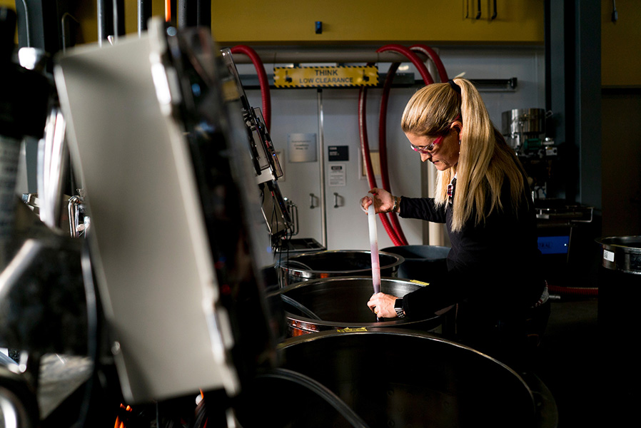 Photograph of a woman leaning over a vat, taking a sample of what looks like red wine.