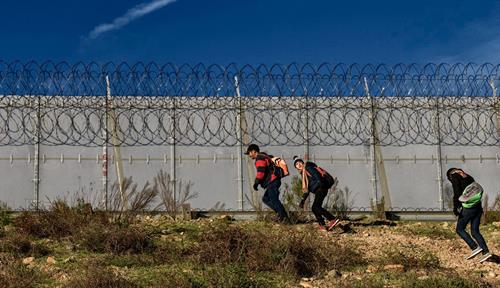 Three people with small backpacks walk through the desert along the base of a wall topped with razor wire.