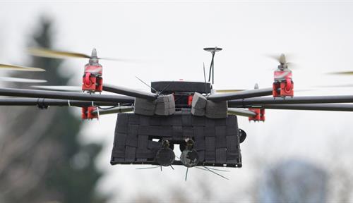 A flying drone with eight rotors flies as three blurry people in the background look on.