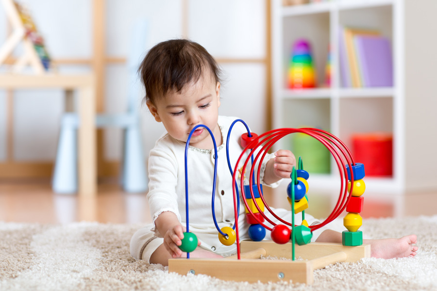 Photograph of a baby in a white onesie sitting on a shag rug playing with a multicolored educational toy.