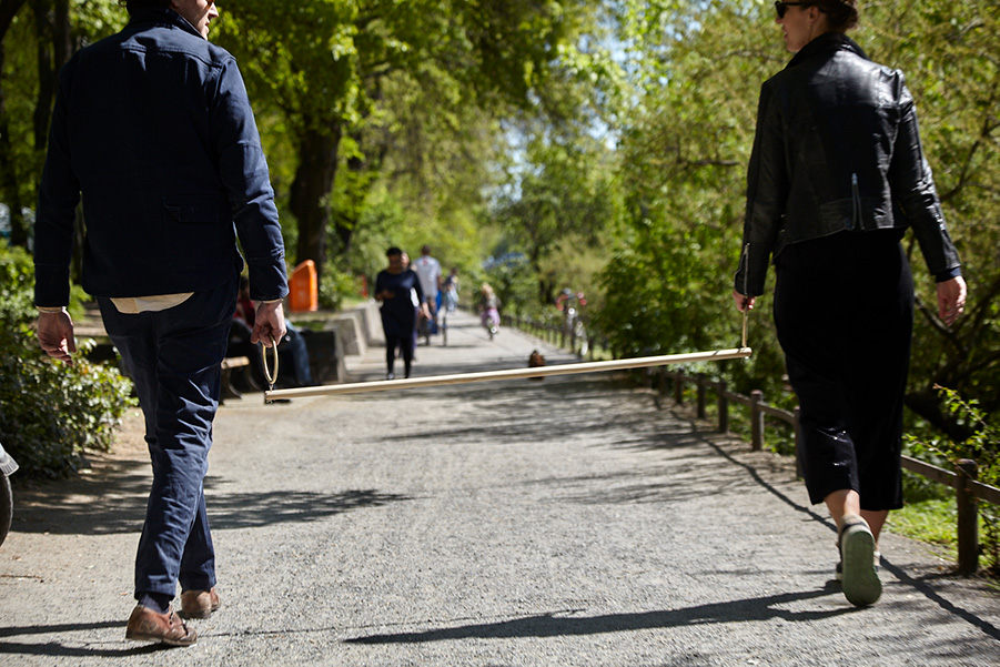 Photograph of two people walking, each one holding a brass ring attached to the ends of a long walking stick.