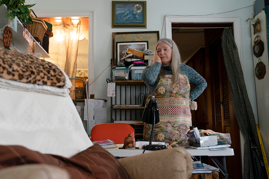 Photograph of a woman with gray hair wearing an apron. She is standing in front of a desk in her home and has one hand held up to her face.