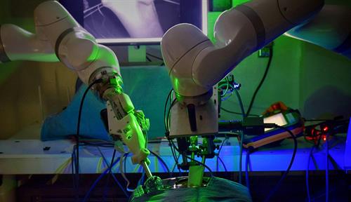 Two robotic arms hover over a draped form. In background a monitor shows a length of intestine gripped by clamps.