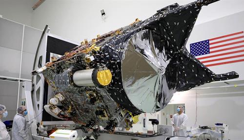 Workers dressed in white clean suits stand around the chassis of the Psyche spacecraft in a facility adorned with a large American flag.