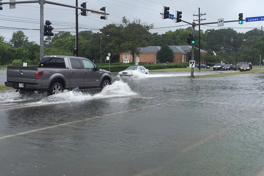 A gray truck and pale car barrel through a heavily flooded street, throwing up spray.