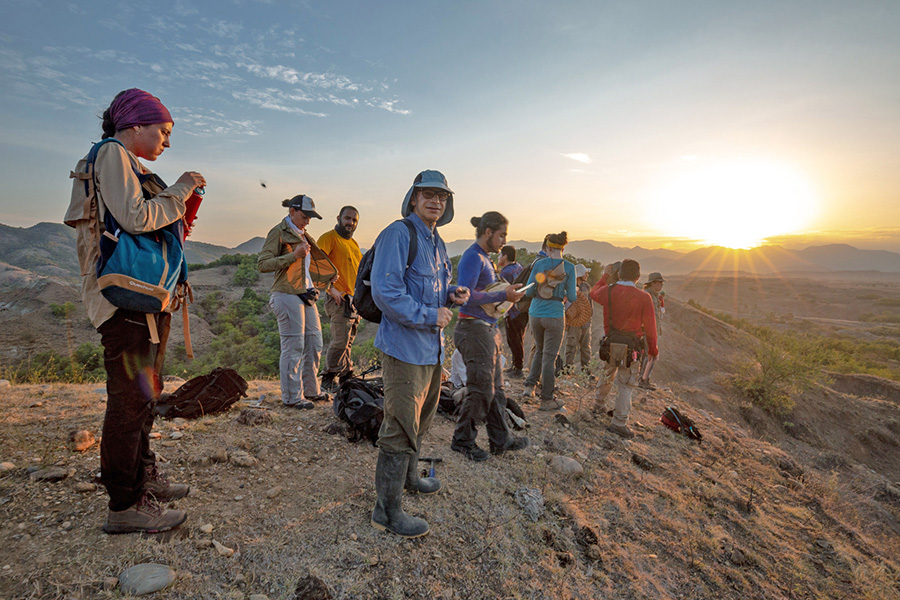 A group of people stand on an arid hill. A desert landscape and the sun shining in the distance can be seen in the background.