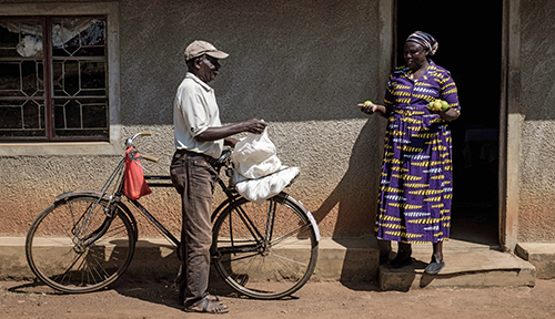 A participant in an experiment on giving people a long-term universal basic income stands outside her house holding oranges she has bought from a man leaning on bicycle in Kenya. 