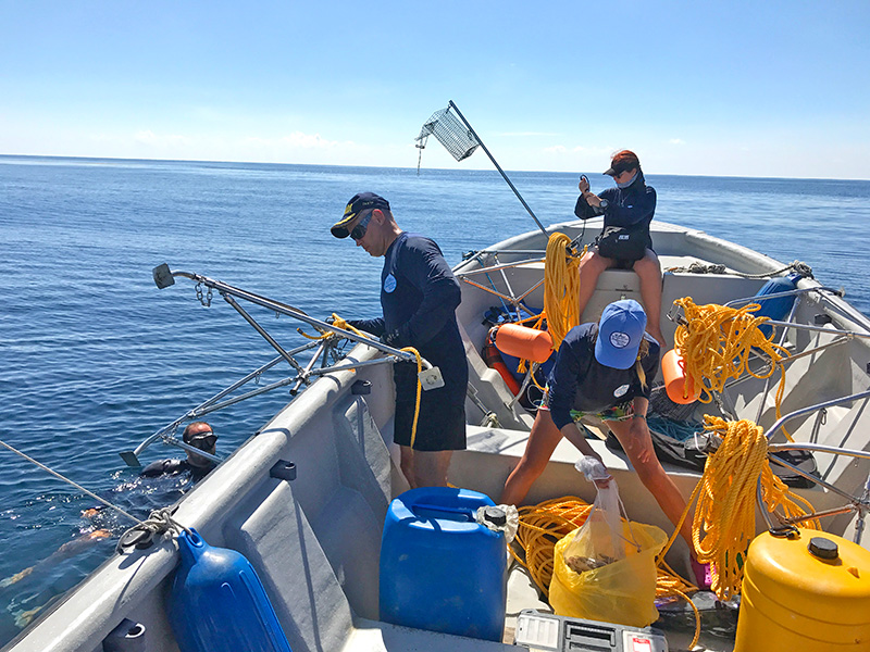 Out in the ocean, three people are aboard a boat and another person is in the water. On the boat, there are blue and yellow containers, as well as cables and other materials.