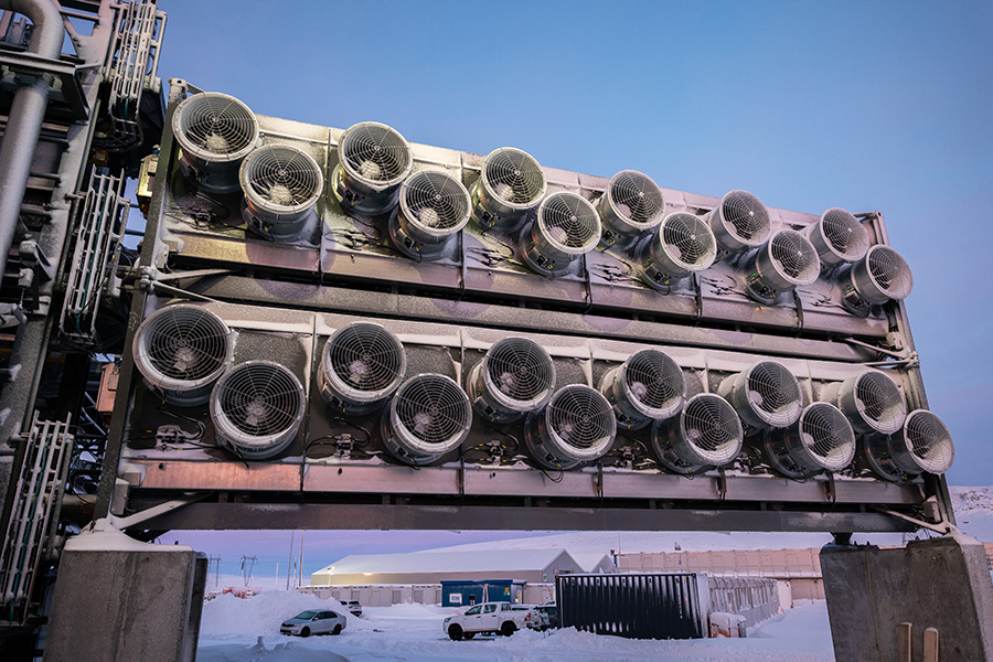 Photo shows a row of a dozen circular ventilators on each of two metal carbon-dioxide-collector containers with blue sky and a snowy parking lot and buildings in the background.