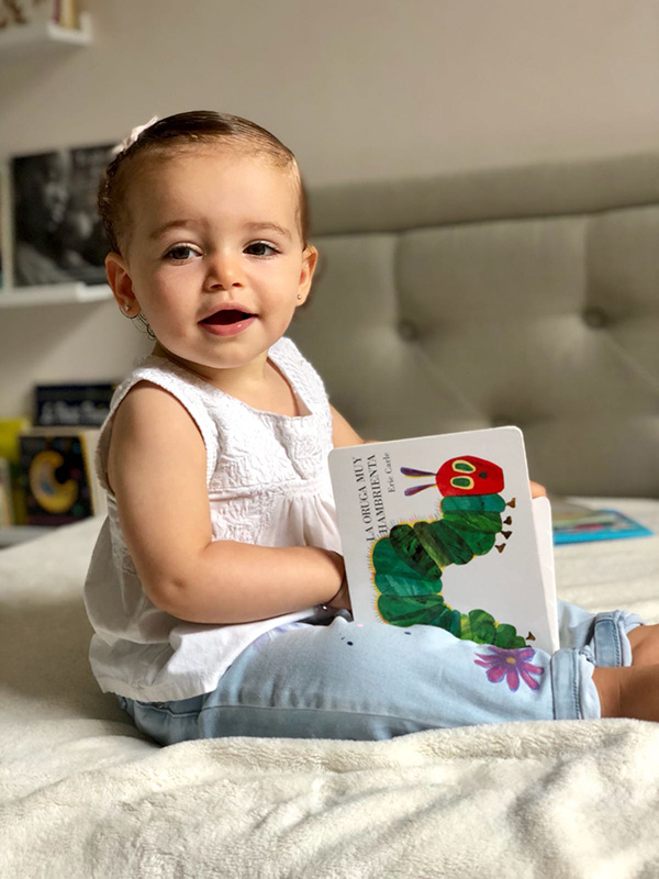 A toddler sits on a bed and holds a book with an illustration of a caterpillar.