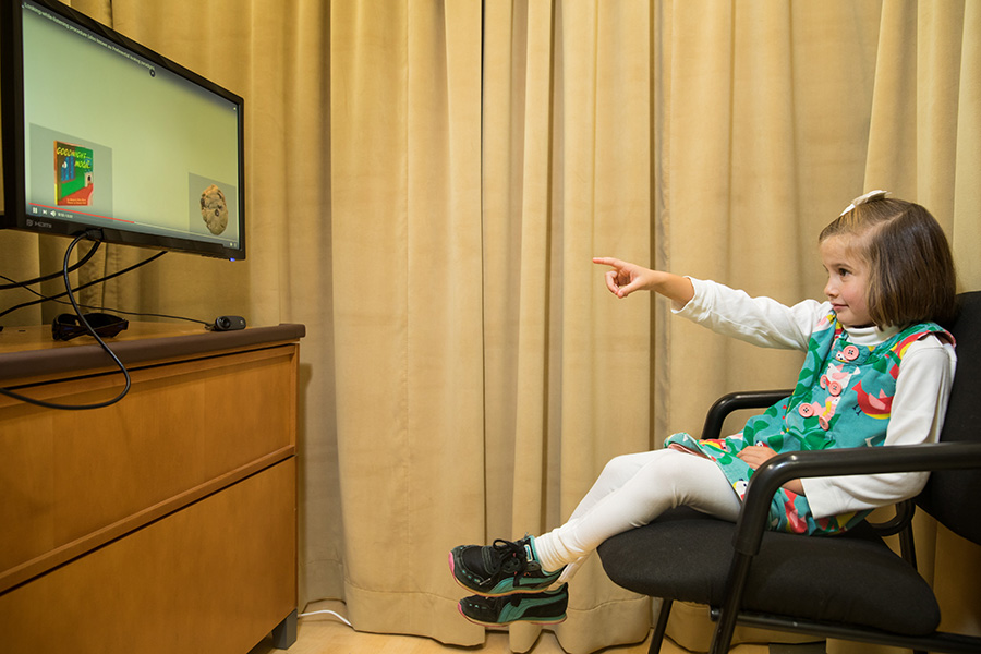 A girl sits in an office chair and points at images she is seeing in a monitor in front of her.