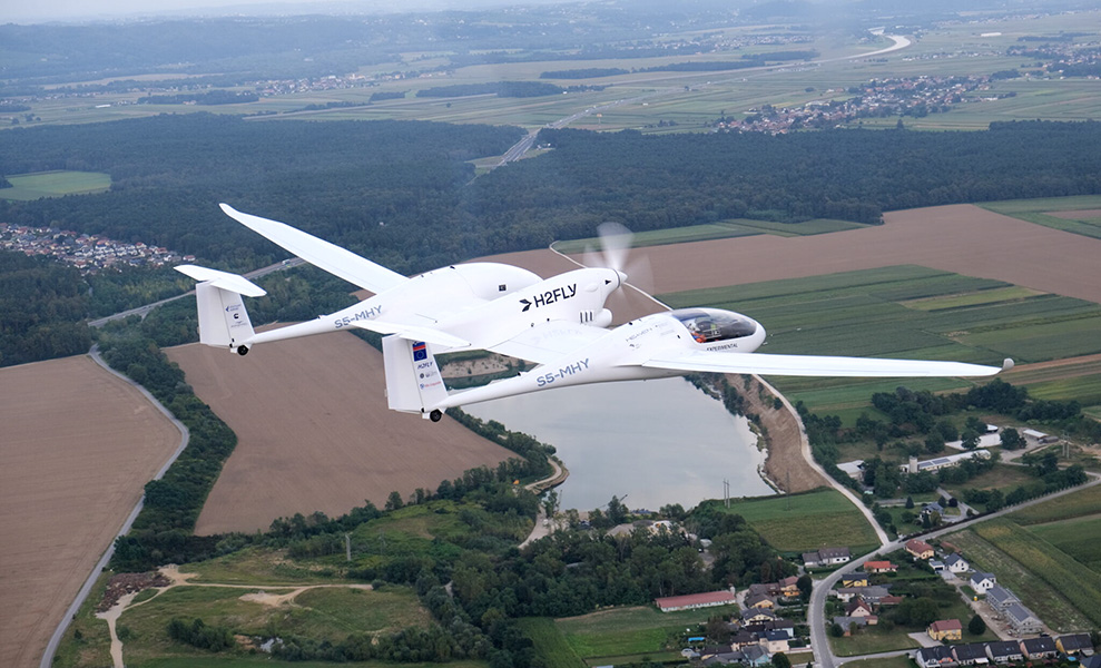  A small white plane with “H2FLY” inscribed on its side flies above fields and houses.