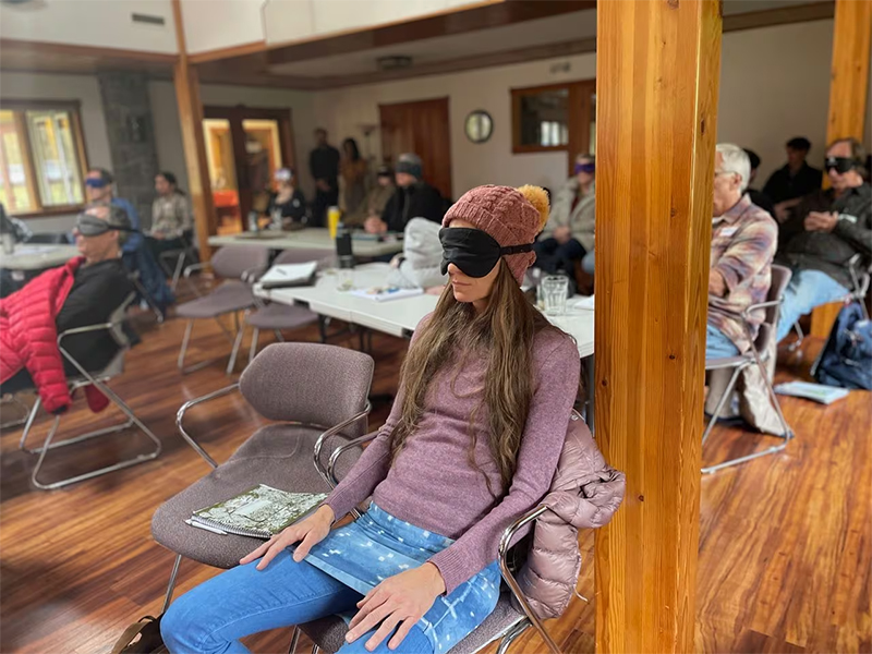 Photo shows a woman wearing a sleeping mask, sitting in a chair in meeting room with others also wearing sleeping masks, all looking toward the front of the room.