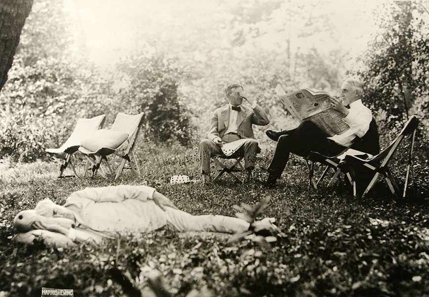 A historic photograph shows a man in white suit sleeping on the lawn while two other men sit in deck chairs behind him reading newspapers.